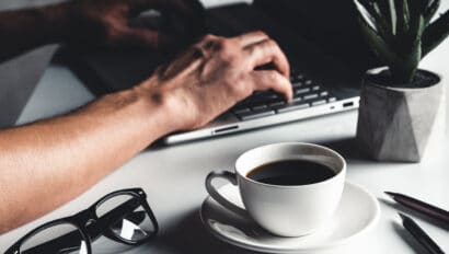 a-man-types-on-a-laptop-business-concept-glasses-a-cup-of-coffee-and-a-pen-on-a-gray-background